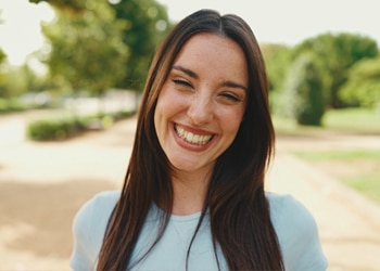 Smiling woman with long dark hair outdoors in light blue shirt, park setting background.
